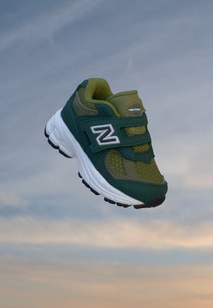 Baskets New Balance vertes et bleu sarcelle foncé avec semelle blanche et bandes velcro flottant sur un fond de ciel nuageux.