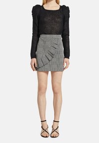 Černý průhledný top s dlouhým rukávem a čtvercovým výstřihem; šedá kostkovaná sukně s volánem; černé sandály na pásky.