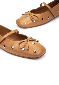 Bottines plates en cuir tan avec un bout rond, un détail en nœud et des clous argentés le long du côté. Dispose d'une fine lanière sur le coup de pied.