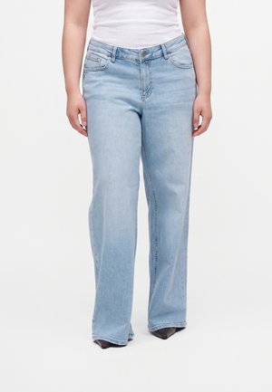 Person iført lyseblå brede jeans og en hvid tætsiddende skjorte, stående med fødderne let adskilt på hvid baggrund.