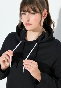 Felpa nera con cappuccio, dotata di una tasca frontale, lacci bianchi con estremità metalliche e una texture morbida e liscia. Vestibilità comoda.