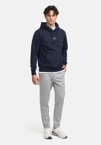 Haut à capuche bleu marine avec poche kangourou et cordons de serrage, associé à un pantalon de survêtement gris clair et des baskets. Design simple avec un détail de logo sur le haut à capuche.