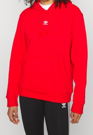 Person iført en knaldrød Adidas hoodie med hvidt logo og sorte Adidas leggings med hvide striber og logo, stående mod en ensfarvet baggrund.