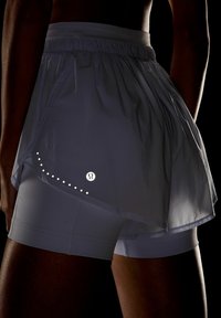 Pantalones cortos de running ligeros, de color gris oscuro, con una superposición reflectante en capas, que presentan un logotipo y acentos en forma de puntos en el costado.