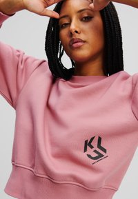 Felpa rosa con un design corto, realizzata in tessuto morbido. Presenta un logo nero sulla parte anteriore e cuciture visibili lungo l'orlo.