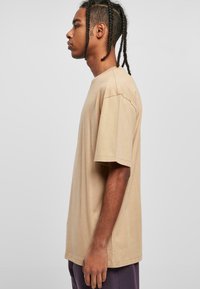 Beige übergroßes Baumwoll-T-Shirt mit kurzen Ärmeln, Rundhalsausschnitt und lässiger Passform, präsentiert an einem Model im Profil vor einem hellen Hintergrund.