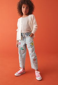 Camiseta gris claro debajo de un cárdigan blanco con canalé; jeans azul claros con bordado floral en varios colores; zapatillas rosa con suelas blancas.