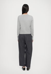 Una donna con lunghi capelli scuri indossa un maglione grigio chiaro, pantaloni larghi grigio scuro con tasche posteriori e scarpe nere con borchie, in piedi contro un muro bianco.