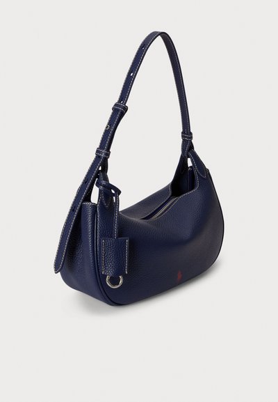 Temno modra usnjena torbica z gladko teksturo, ukrivljeno obliko in nastavljivim trakom. Ima zadrgo in majhno etiketo z kovinskim obročkom.