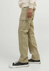 Beige cargo-byxor med avslappnad passform, featuring flera fickor, sömnadsdetaljer och använda med svarta sneakers som har en kontrasterande vit sula.