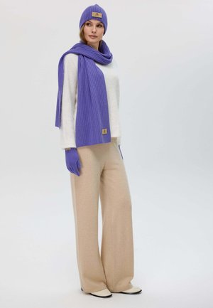 Cappello a coste viola, sciarpa e guanti abbinati a un maglione bianco e pantaloni beige a gamba larga. Materiali testurizzati con dettagli logo.