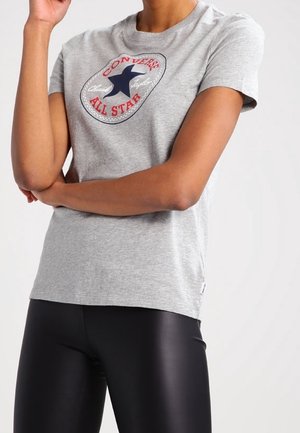 Persoon draagt een grijs Converse All Star T-shirt en zwarte glanzende legging, met gekruiste armen en hand dichtbij het gezicht.