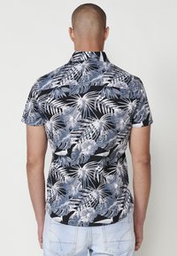 Camicia a maniche corte con sfondo nero e motivo di foglie tropicali bianche e blu chiaro. Il tessuto sembra leggero e traspirante.