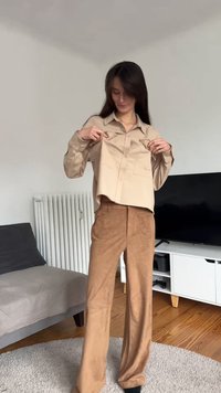 Beige overhemd met knopen en voorzakken, gecombineerd met bruine wijde suède broek. De outfit wordt gemodelleerd in een minimalistische binnenomgeving.