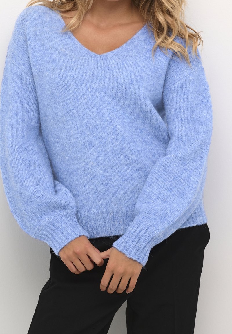 Femme portant un pull en maille bleu clair à manches longues et un pantalon noir sur un fond uni.
