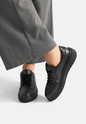 Osoba nosząca czarne skórzane trampki sznurowane z grubymi podeszwami i szare szerokie spodnie na białym tle.