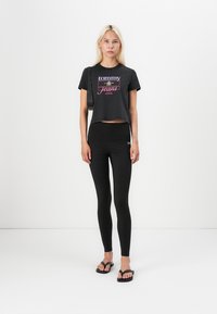 Juodas trumpas marškinėliai su "Tommy Jeans" logotipu, derinami su aukšto liemens juodomis leginsais ir juodais šlepetėmis, pateikti ant baltos fono.