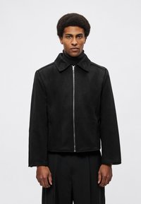 Crna flis jakna s prednjim zatvaračem na patent, šiljastim ovratnikom i širokim rukavima. Ima detalje šivanja i teksturirani naglasak na ovratniku.