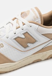 Białe i beżowe sneakersy z wyściółką wokół kostki, perforowanymi detalami oraz wyraźnym wytłoczonym logo. Posiadają płaskie sznurowadła i teksturowaną podeszwę.