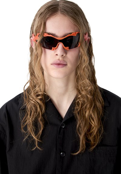Person mit langen, lockigen Haaren trägt ein schwarzes Hemd und futuristische schwarze Sonnenbrille mit auffälligen orangefarbenen Rahmen.