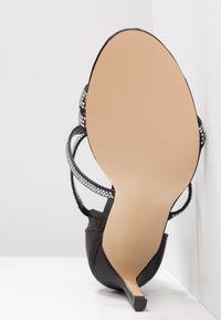 Chaussure à talons hauts noire avec une surface lisse, un dos ouvert et des brides ornées de strass. La semelle est de couleur claire et texturée.