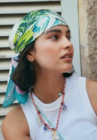 Zijden sjaal met groene en blauwe botanische print; gedragen met een witte mouwloze top, gelaagde kettingen en schouderlang donker haar.