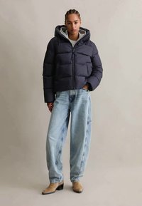 Kvinde iført en marineblå puffjakke med hætte, lyseblå vidde jeans og brune ankelstøvler, stående foran en ensfarvet baggrund.