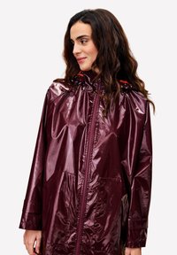 Bordeaux waterdichte regenkap met een glanzende afwerking, rits aan de voorkant, verstelbare capuchon en zijzakken. Heeft lange mouwen en een losse fit.