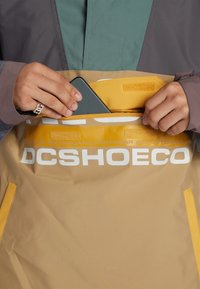 Gelbe Jacke mit grauen und grünen Akzenten, die eine Reißverschlusstasche hat. Sichtbares Logo "DCSHOECO" auf der Vorderseite. Nahaufnahme eines Handys, das eingeführt wird.