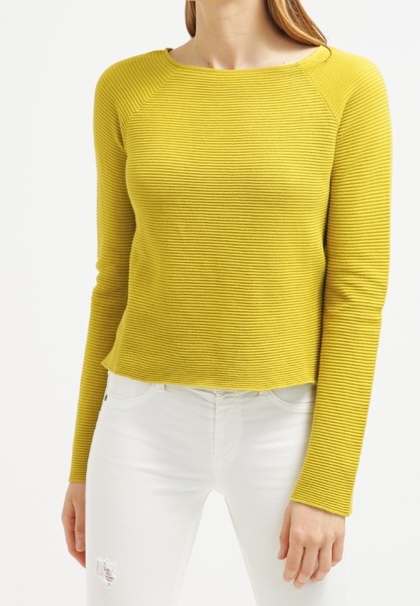 Pull à manches longues côtelé jaune avec un col rond, présentant un tissu texturé. Associé à un jean blanc déchiré.