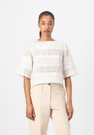 Crèmekleurige blouse met korte mouwen, voorzien van gestructureerde strepen en bloemvormige uitsparingen; gecombineerd met beige broeken met een knoopsluiting.