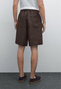 Shorts en lin marron avec une taille élastique et deux poches arrière, assortis à des chaussures foncées sur une surface texturée et sombre.