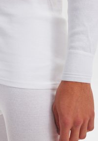 Hvid langærmet top med ribbet tekstur og manchetter, parret med hvide bukser. Glat stof med subtile syninger.