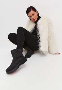 Botas negras de tobillo insulated con un diseño acolchado, que cuentan con un acento de botón redondo y suela texturizada, llevadas con leggings negros y un abrigo de piel sintética blanco.