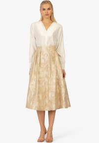 Jupe beige à broderies florales avec taille haute et plis, assortie à un chemisier blanc à manches longues. Sandales à talons transparents.