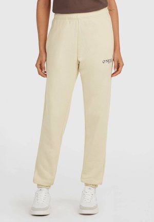 Persoon draagt beige joggingbroek met elastische enkels, een klein O'Neill-logo op de bovenste linker dij en witte sneakers met grijze accenten.