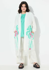 Pitkä valkoinen kimono, jossa on värikäs vaaleanpunainen ja vihreä kukkakuosi, jossa on frangeerausreunat. Yhdistetty turkoosinvärisen paidan ja valkoisten housujen kanssa.