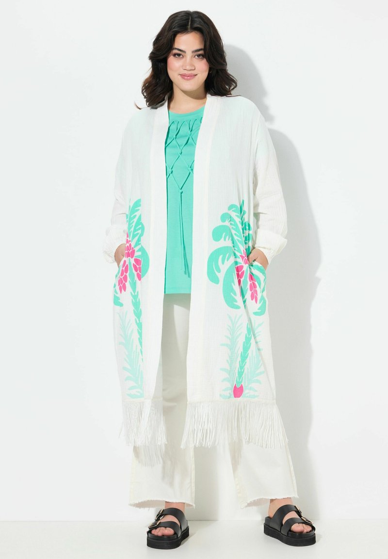 Lång vit kimono med färgglad blommig tryck i rosa och grönt, med fransar längs kanterna. Kombineras med en turkos topp och vita byxor.