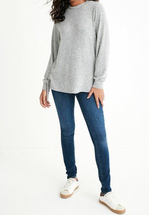 Femme portant un pull léger à manches longues gris clair, un jean skinny bleu foncé et des baskets blanches, debout devant un fond blanc.