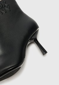 Bottine en cuir noir avec un design épuré, bout pointu et un talon haut brillant. Présente un détail de logo embossé sur le côté.