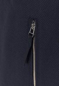 Marinblå textil med struktur som har en dragkedja med metallicdragare och svarta accenter. Ytan visar ett rutmönster.