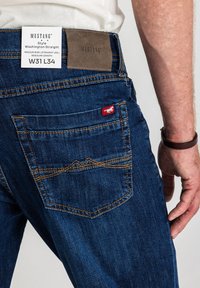 Niebieskie dżinsy o średnim stanie i prostych nogawkach. Charakteryzują się pomarańczowym przeszyciem, brązową skórzaną naszywką oraz czerwoną metką na kieszeni.