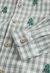 Chemise en flanelle à carreaux verts et blancs avec des boutons en bois et des arbres verts brodés. Texture douce, poignet à bouton et détails du col visibles.