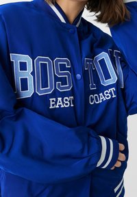 Modrá bomber bunda s bílým vyšitým textem "BOSTON EAST COAST", druky a proužkovanými manžetami. Hladká, lehká látka.
