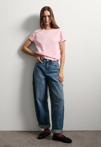 Camiseta de gran tamaño color rosa claro, jeans de mezclilla azul de tiro alto con puños enrollados, combinados con mocasines negros y calcetines rojos, de pie contra una pared.