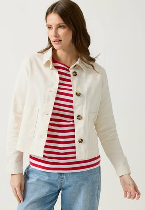 Donna che indossa una giacca crema con bottoni sopra una camicia a righe rosse e bianche con jeans azzurri chiari su sfondo neutro.