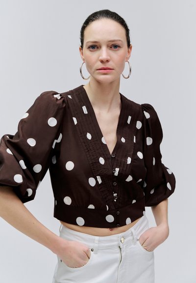 Tumejuukseline naine hõbekõrvarõngastega, kandes pruuni lühikest pluusi valgete täppidega ja valgeid pükse, käed taskutes, neutraalne taust.