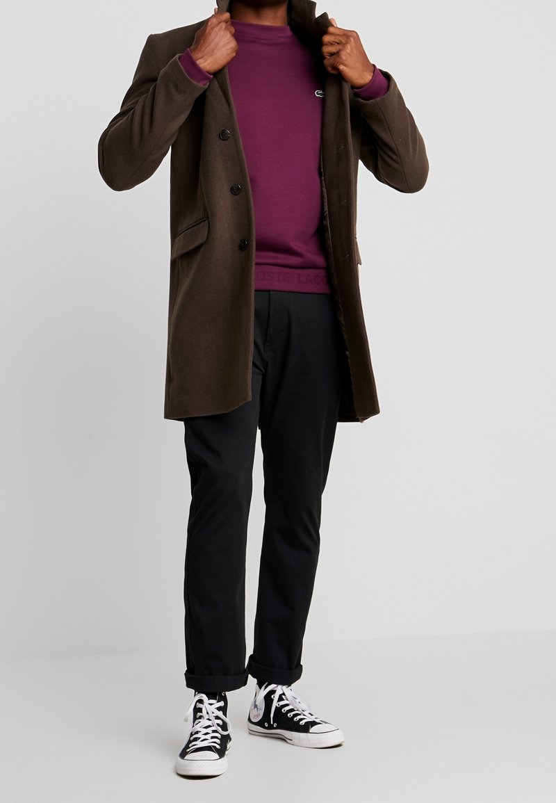 Homme portant un manteau marron, un sweat-shirt violet, un pantalon noir retroussé et des baskets montantes noires ajustant le col de son manteau.