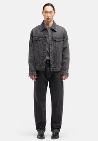 Schwarze Jeansjacke mit zwei Brusttaschen, kombiniert mit schwarzer Jeans. Das Model trägt ein graues T-Shirt darunter und schwarze Lederstiefel.