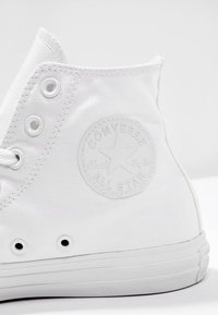 Baskets montantes en toile blanche avec un patch logo rond en relief, des œillets métalliques et une semelle en caoutchouc texturée.